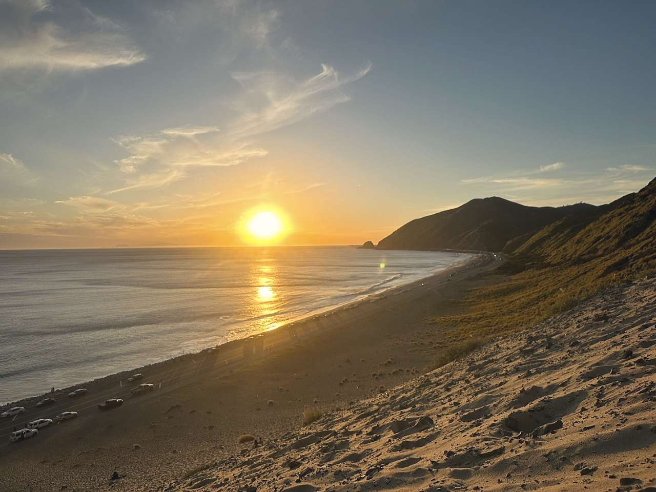 The sun low over the water, a long beach and a road running toward dark hills.